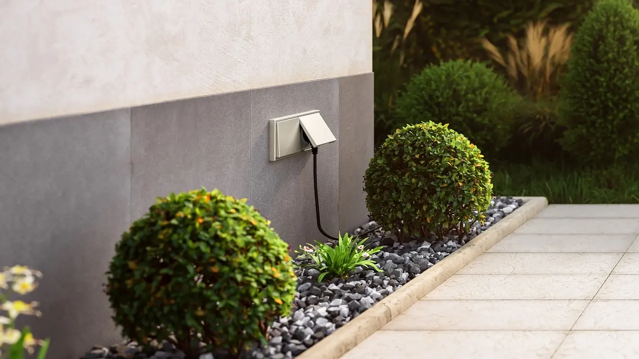 Prise de jardin étanche à clapet encastrée dans un mur extérieur, câble noir branché, en bordure d'une terrasse carrelée