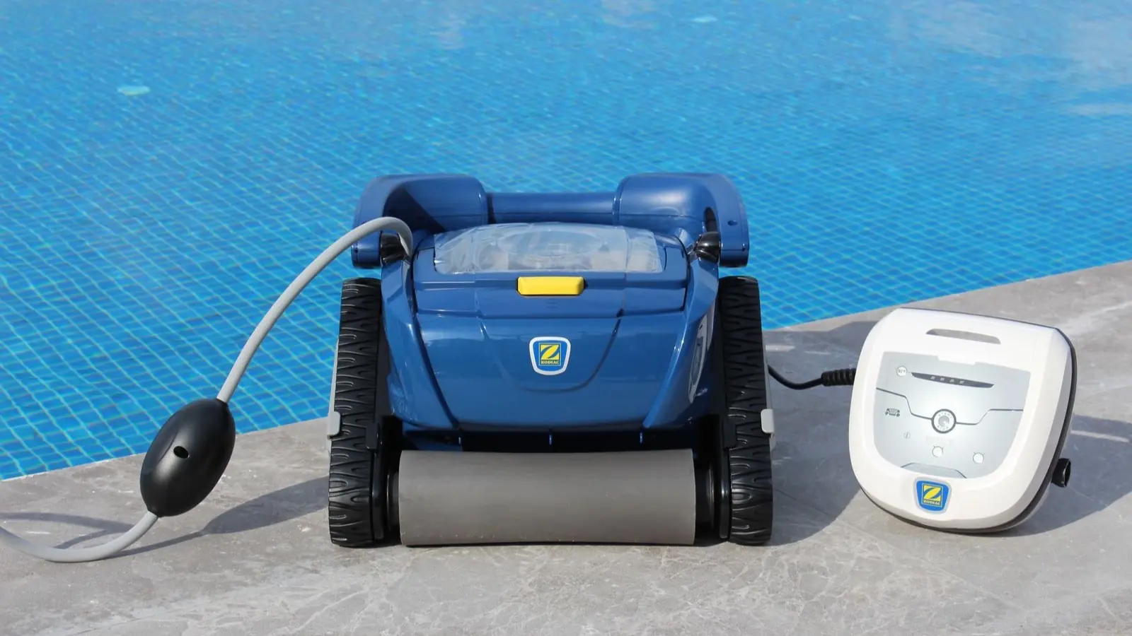 Robot de piscine électrique bleu à chenilles posé sur la margelle à côté de son bloc d'alimentation blanc, câble et flotteur déroulés devant le bassin