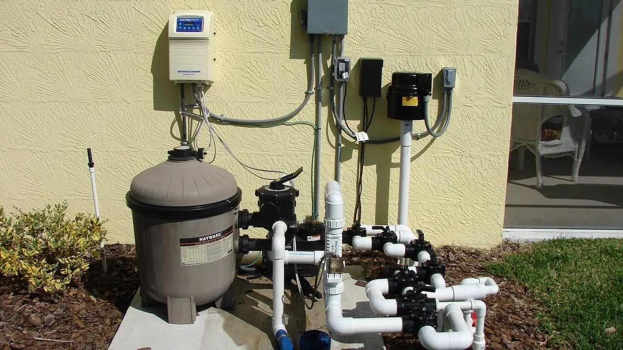 Local technique de piscine en extérieur : filtre à sable, pompe de filtration, second moteur électrique et coffrets de commande reliés par des tuyaux PVC blancs