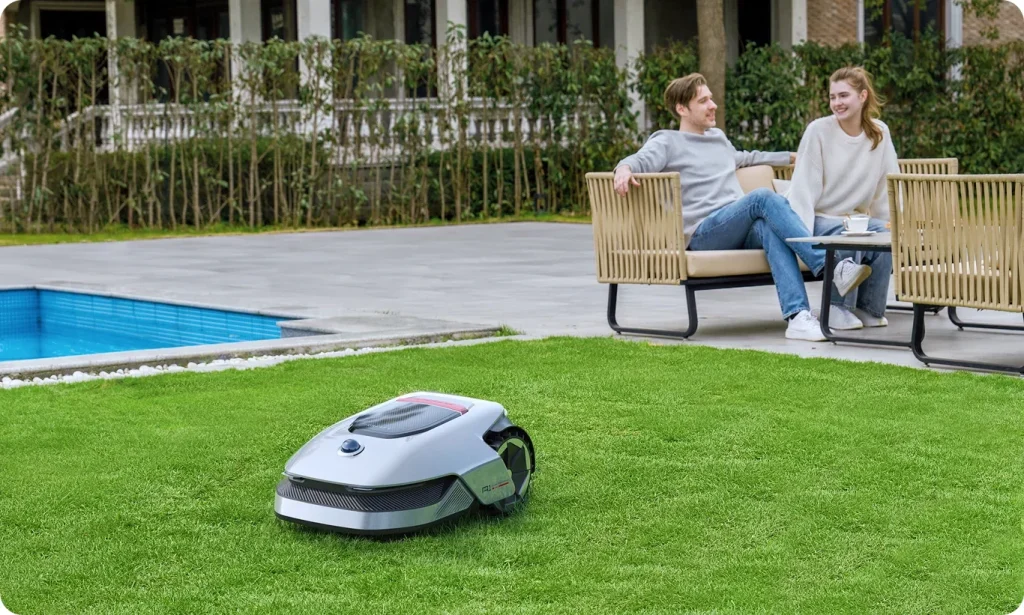 Robot tondeuse Dreame A1 tondant la pelouse au bord d'une piscine pendant qu'un couple discute sur la terrasse