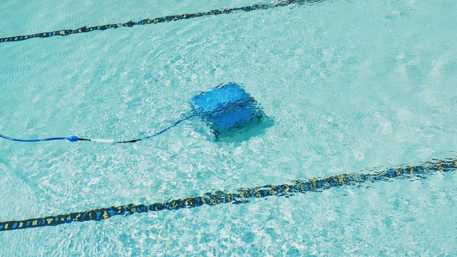 Est-ce qu'un robot de piscine abîme le liner : robot nettoyeur bleu relié à son tuyau, posé au fond d'un bassin turquoise sous les lignes d'eau