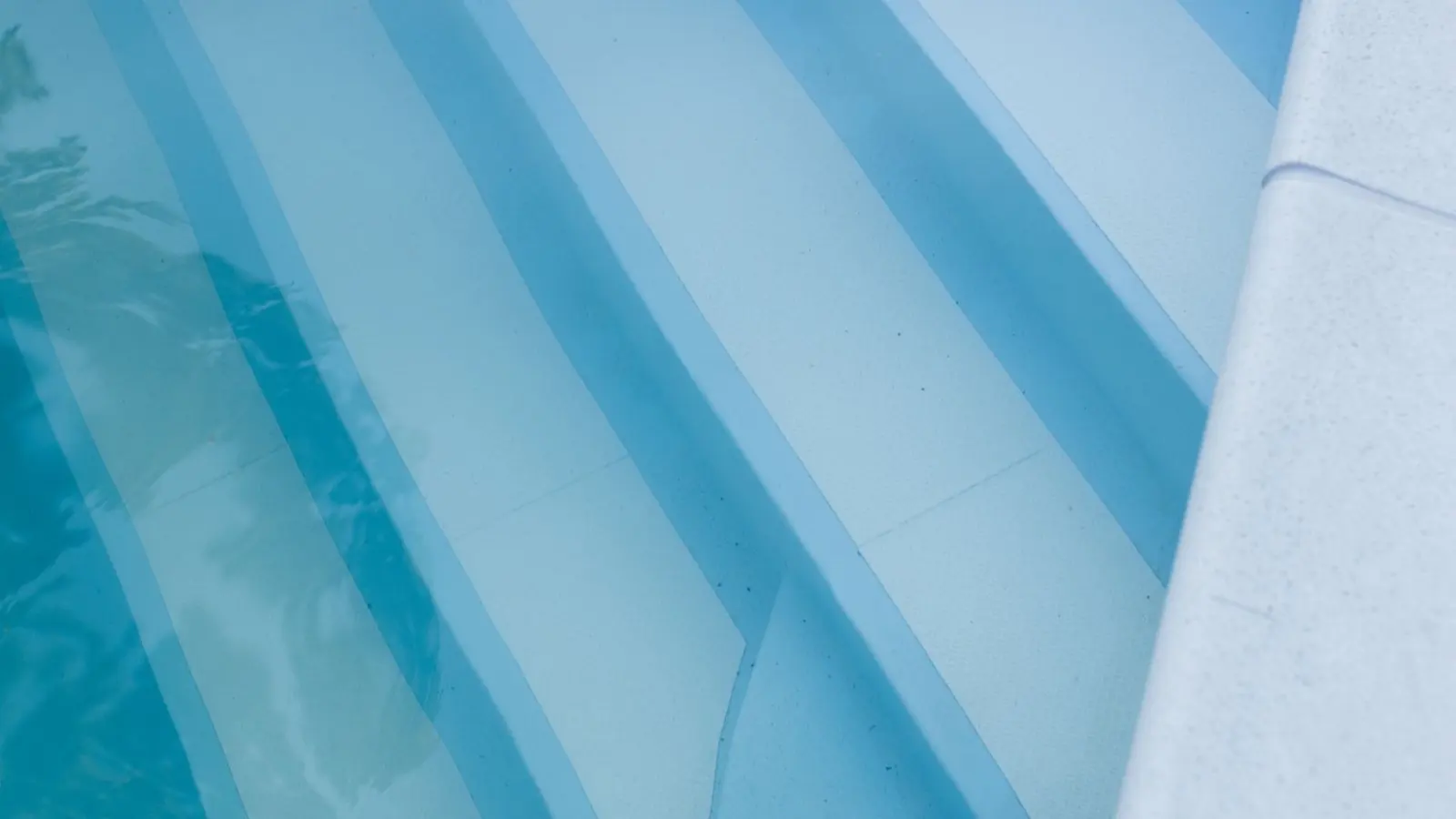 Escalier immergé d'une piscine recouverte d'un liner bleu clair, marches et nez de marche vus à travers l'eau depuis la margelle blanche