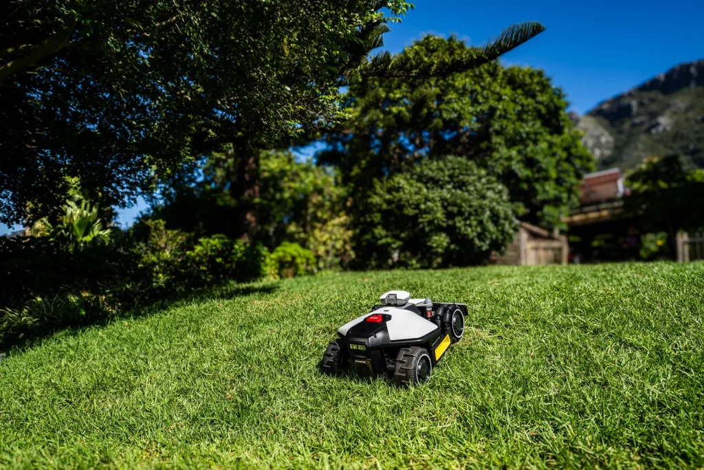 Robot tondeuse Mammotion Luba Mini en train de tondre une pelouse vallonnée bordée d'arbres, vu de trois quarts arrière