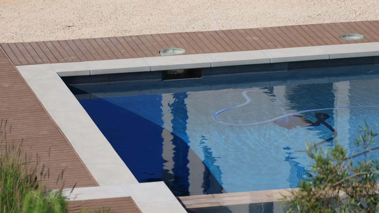 Vue plongeante sur un bassin rectangulaire bordé d'une terrasse en bois : le tuyau souple d'un nettoyeur de piscine serpente dans l'eau jusqu'à l'appareil immergé près du bord