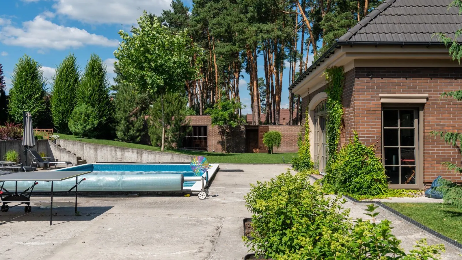 Piscine rectangulaire enterrée dans un jardin arboré, avec sa couverture à bulles à demi enroulée sur un enrouleur mobile posé au bord du bassin.