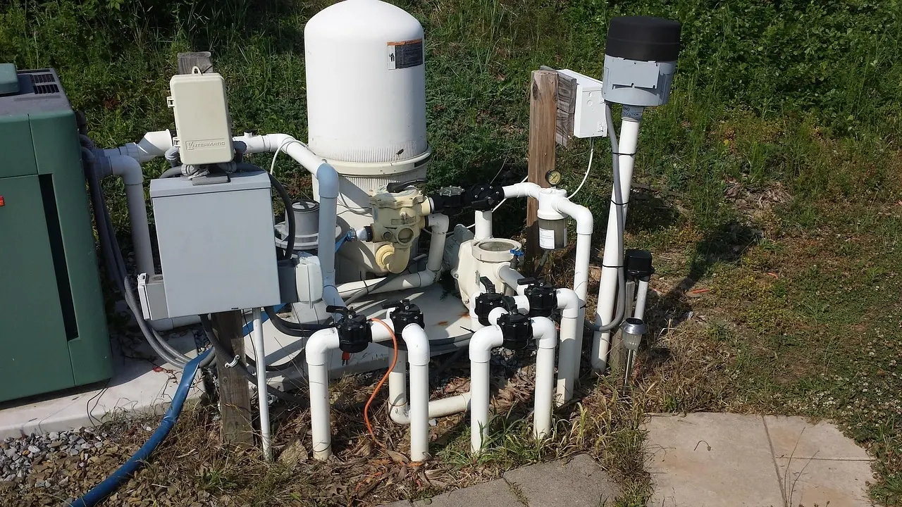 Installation de filtration de piscine à l'extérieur : cuve de filtre blanche, pompe, vannes noires et réseau de tuyaux PVC reliés à des coffrets électriques.