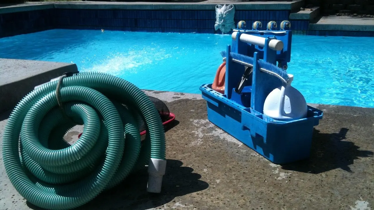 Matériel manuel d'entretien de piscine posé sur la margelle : tuyau d'aspiration enroulé, brosses et tête d'aspirateur dans un bac bleu